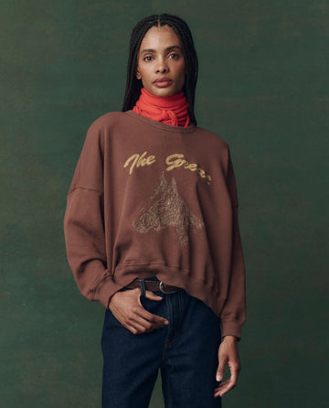 A person in THE GREAT. F25 D2’s The Teammate Sweatshirt (Umber, horse embroidery), a red scarf, and dark jeans stands against a green background.