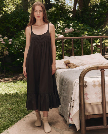 A woman in THE GREAT. SP25 SLEEP The Eyelet Ruffle Tank Night Dress in black, featuring Victorian-inspired eyelet lace trim, stands by a vintage bed with a quilt and teddy bear in a garden setting.