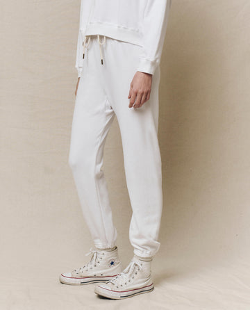 An individual wearing The Stadium Sweatpant in True White by THE GREAT. HOL 24 KNITS, paired with vintage high-top sneakers, stands against a beige background.