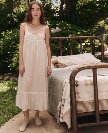 A woman in THE GREAT. SP25 SLEEP's Eyelet Ruffle Tank Night Dress in True White stands barefoot by an old-fashioned bed outdoors, surrounded by greenery and flowers. A teddy bear sits on the bed.
