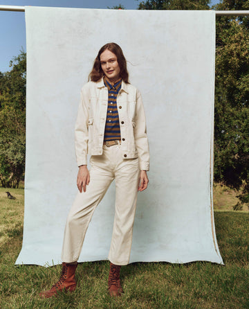 A person in a white denim set stands outdoors, wearing THE GREAT. PS25's The Workwear Jean Jacket in Natural Hemp over a striped shirt, paired with brown boots. The natural backdrop of trees and grass enhances this timeless style mixing casual elegance with classic workwear vibes.