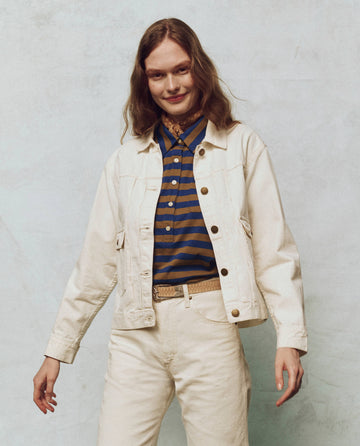 Someone wearing The Workwear Jean Jacket in Natural Hemp from THE GREAT. PS25 over a striped shirt and beige pants stands against a textured background.