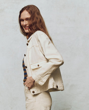 A person smiling, in THE GREAT. PS25's The Workwear Jean Jacket in Natural Hemp, effortlessly exudes style. This look nods to classic workwear aesthetics against a plain, light-colored backdrop.