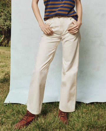 A person in THE GREAT. PS25's Workwear Jean in Natural Hemp stands on the grass, wearing a striped top and brown boots, with a light blue backdrop behind them.