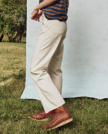 A person in The Workwear Jean by THE GREAT. PS25 stands on the grass in a striped shirt and beige pants, embodying classic workwear style. Facing a light background, they’re grounded by sturdy brown boots, showcasing an effortless blend of function and fashion.