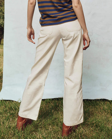 A person in a striped shirt and THE GREAT. PS25's The Workwear Jean in Natural Hemp stands on the grass, facing away from the camera.
