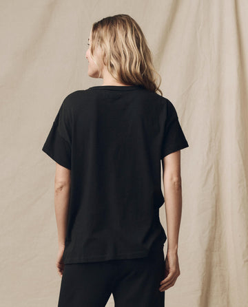 A person with long, wavy hair wearing The Boxy Crew. Solid -- Almost Black t-shirt by THE GREAT. CORE KNITS and black pants stands facing away against a beige background.
