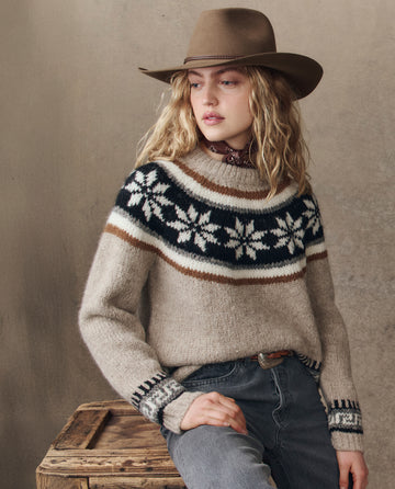 A person wearing THE GREAT. H25 COLLECTION's The Snowflake Fairisle Pullover in Oat, a brown hat, and gray jeans sits on a wooden crate against a neutral background.