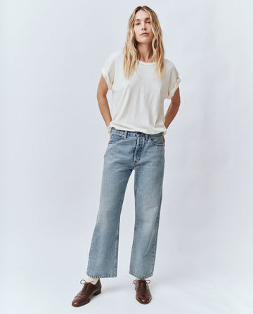 A person with long hair stands against a plain background, wearing a white t-shirt, brown shoes, and The Wayne Jean in Sedona Wash by THE GREAT. PF24.