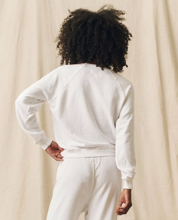 A person with curly hair stands facing away, wearing **The Shrunken Sweatshirt, Solid -- True White** by **THE GREAT. FALL 24 KNITS**, paired with locally handcrafted sweatpants, against a beige curtain background.