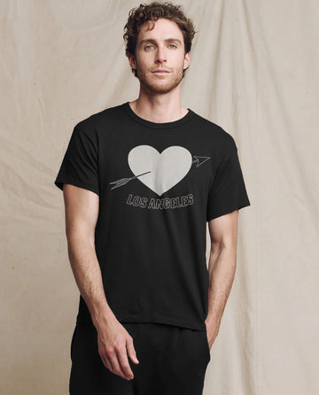A man stands against a plain backdrop in The Men's Boxy Crew by THE GREAT., showcasing an Almost Black LA heart graphic with an arrow, encapsulating Los Angeles cool.