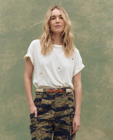 A woman with wavy blonde hair wears THE GREAT. SU25 The Boxy Crew. Embroidered—Washed White with Floating Daisy Embroidery, paired with camouflage pants, standing against a textured green background.