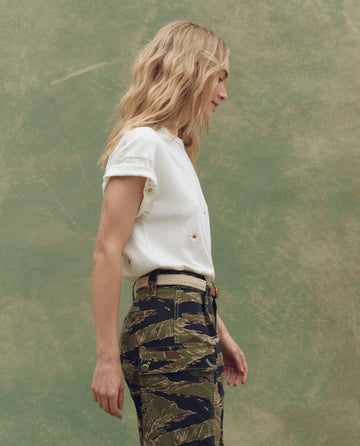 A person with long blonde hair stands in profile against a green textured background, wearing THE GREAT. SU25's The Boxy Crew. Embroidered in Washed White with Floating Daisy Embroidery and camouflage pants.