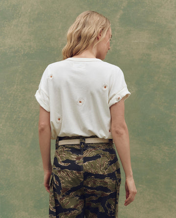 A person with wavy blonde hair, facing away from the camera against a green background, wears THE GREAT. SU25's Boxy Crew. Embroidered tee in washed white with floating daisy embroidery, paired with camouflage pants.