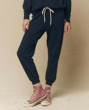 A person wearing black sweatpants, a dark sweatshirt, and pink high-top sneakers with white laces stands against a beige background, showcasing the comfort of The Cropped Sweatpant. Solid -- True Navy by THE GREAT. FALL 23 KNITS, made from 100% cotton vintage gym pants.