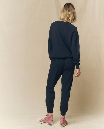 A person with shoulder-length hair, wearing a dark sweatshirt, THE GREAT.'s Cropped Sweatpants in True Navy from the FALL 23 KNITS collection, and pink high-top sneakers stands facing away from the camera against a beige background.