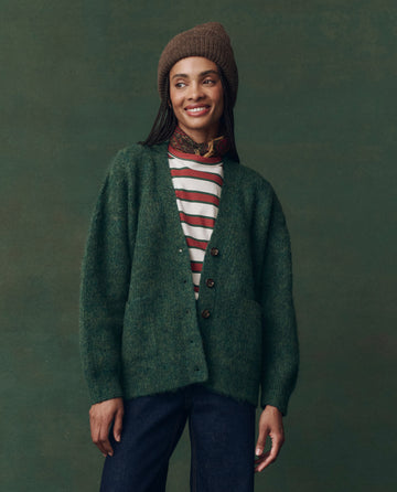 Wearing THE GREAT. F25 D2’s Brushed Melange Cardigan in Meadow Green, a person stands against a green background, in a striped shirt, brown knit hat, and dark jeans, smiling and glancing to the side.