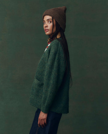 A person in THE GREAT. F25 D2's Brushed Melange Cardigan in Meadow Green, paired with a brown knit hat and dark pants, stands sideways against a plain green background.