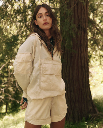 Wearing a beige hoodie and THE GREAT. SP25 TGO's The Rover Short in Cream, this individual embodies effortless casual wear outdoors by a tree.