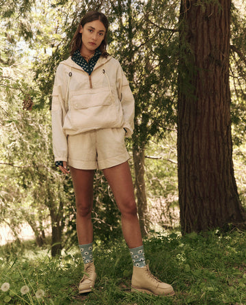 A woman stands in a wooded area wearing The Rover Short in Cream by THE GREAT. SP25 TGO, a beige jacket, blue patterned socks, and tan boots, with trees and grass surrounding her.