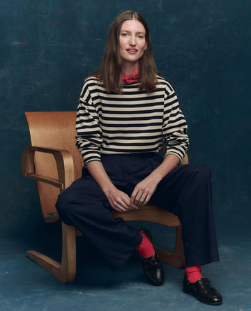 A person sits on a wooden chair against a blue background, wearing THE GREAT. SP25 COLLECTORS The Sailor Sweater -- Black Stripe, dark pants, red socks, black shoes, and a red scarf around their neck.