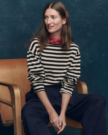 A woman sits on a wooden chair against a dark blue background, wearing THE GREAT. SP25 COLLECTORS' The Sailor Sweater in Black Stripe, paired with dark pants and a red bandana around her neck.