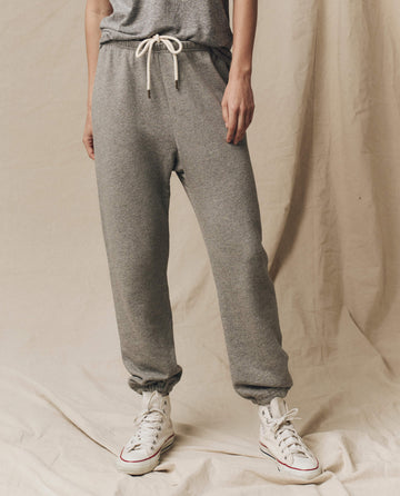 A person wearing The Stadium Sweatpant in Varsity Grey by THE GREAT. CORE KNITS and white sneakers stands against a beige fabric backdrop. The vintage athletic sweatpants feature an elasticated waist and gathered ankles.