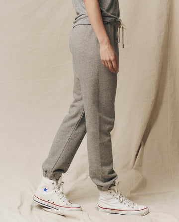 A person in a gray outfit, featuring The Stadium Sweatpant in Varsity Grey by THE GREAT. CORE KNITS with an elasticated waist, and white high-top Converse sneakers stands facing sideways against a beige backdrop.