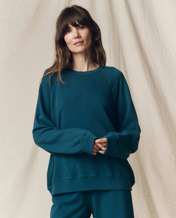 A woman with long brown hair wears THE GREAT.’s The Slouch Sweatshirt in Solid Dark Slate from the F25 SEASONAL KNITS collection, paired with matching pants, standing before a neutral beige fabric backdrop.