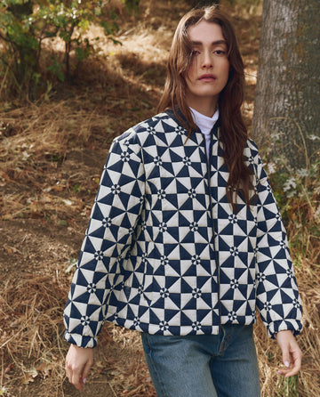 A person stands outdoors in THE GREAT. F25 TGO The Alps Puffer Jacket—Quilted Vintage Navy—paired with a white turtleneck and blue jeans, surrounded by dry grass and trees.