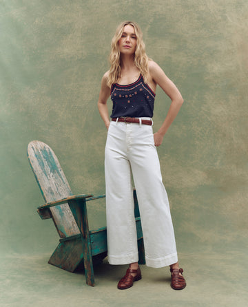 A woman stands beside a weathered wooden chair, wearing THE GREAT. SP25 D1 The Seafair Jean in True White, a dark sleeveless top, and brown shoes against a green textured backdrop.