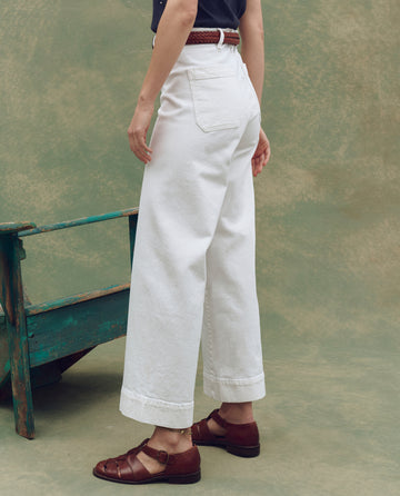 The lower body is shown standing in front of a greenish background, wearing THE GREAT. SP25 D1 The Seafair Jean in True White, styled with a brown woven belt and brown sandals.
