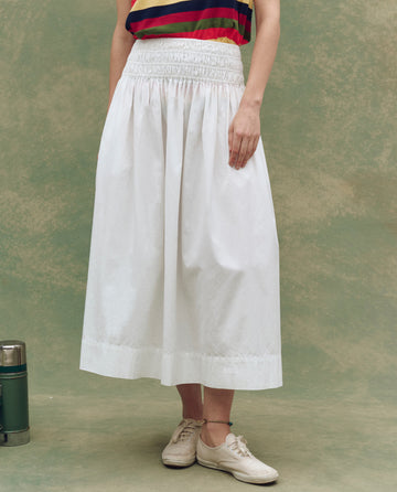 A person in THE GREAT. SU25’s white "The Viola Skirt," paired with a striped sleeveless top, white shoes, and anklet stands before a green textured backdrop—an elegant women’s fashion look.