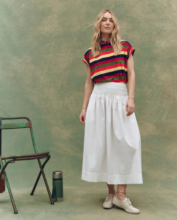 A woman poses before a mottled green backdrop in a striped top, THE GREAT. SU25 The Viola Skirt in White, and beige shoes; beside her are a green metal chair and thermos, showcasing chic women’s fashion.
