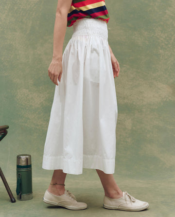 A person stands sideways wearing a sleeveless striped top, The Viola Skirt in white by THE GREAT. SU25, white shoes, and an anklet. A thermos and chair in the background highlight chic women’s fashion.
