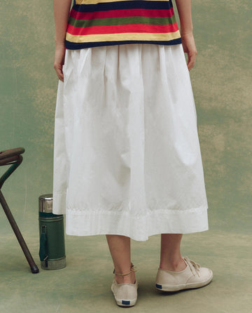 A person stands facing away, modeling THE GREAT. SU25's The Viola Skirt in White with a striped top and beige shoes. A green thermos and part of a chair are beside them, set against a green background.