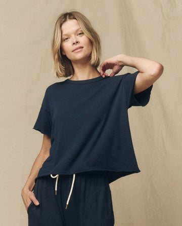 A person with shoulder-length blonde hair wearing THE GREAT. FALL 23 KNITS' "The Crop Tee. Solid -- True Navy" and matching high-rise jeans poses in front of a plain beige background.
