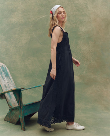 A woman in THE GREAT. SU25 The Attic Lace Dress in black, white sneakers, and a headscarf stands by a weathered green wooden chair against a textured green background.