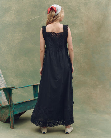 A woman stands facing away in THE GREAT. SU25 The Attic Lace Dress – Black, featuring a long lace-trimmed hem. She wears white shoes and a white headscarf with a red accent. A weathered green bench sits beside her against a neutral background.