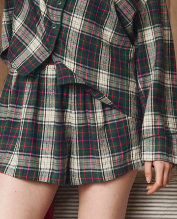 A person models THE GREAT. H25 SLEEP's The Flannel Smocked Sleep Short in Pine Lodge Plaid, showcasing the green, white, and red plaid pattern from shoulders to mid-thigh.