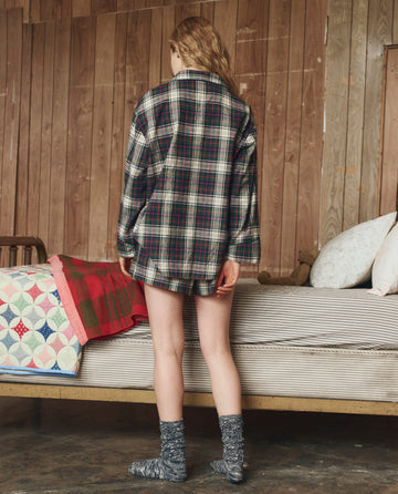 A person stands indoors facing a wooden wall, wearing THE GREAT. H25 SLEEP’s The Flannel Pajama Top in Pine Lodge Plaid and gray socks, with no pants. A bed with a colorful quilt is visible on the left.