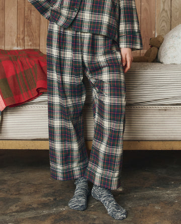 A person wearing THE GREAT. H25 SLEEP The Flannel Easy Sleep Pant in Pine Lodge Plaid stands next to a bed with a striped blanket and red plaid throw, set against a wooden wall background.