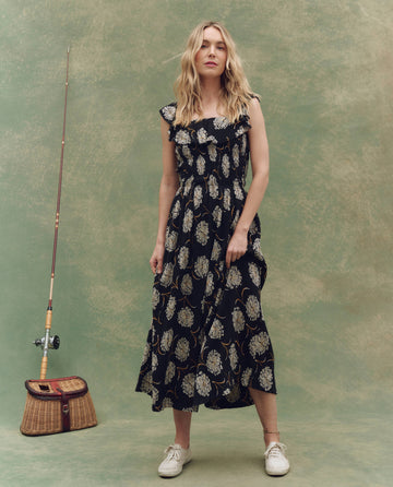 A woman wearing THE GREAT. SU25’s The Havana Dress in Black Dancing Floral with white sneakers stands on a mottled green background beside a fishing rod and wicker basket.