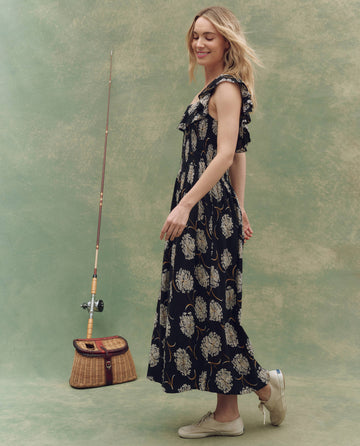 A woman smiles sideways next to a fishing rod and wicker basket, wearing THE GREAT. SU25's The Havana Dress in Black Dancing Floral and white sneakers, set against a muted green background.
