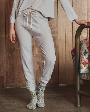 A person indoors wears THE GREAT. H25 SLEEP The Long John in Heather Grey—light gray men's base layer lounge pants and matching long-sleeve top—with gray knit socks, standing by a bed with a colorful quilt.