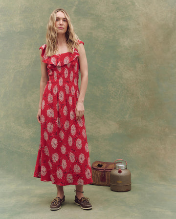 A woman in THE GREAT. SU25's Havana Dress in True Red Dancing Floral stands before a textured backdrop, brown shoes on her feet, with a vintage thermos and woven case nearby, embodying timeless summer style.