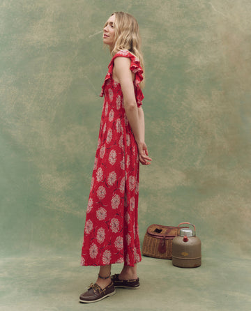 A woman in profile wears THE GREAT. SU25’s The Havana Dress in True Red Dancing Floral, hands behind her back. A wicker basket and metal container rest nearby, all set against a mottled green backdrop for effortless summer elegance.