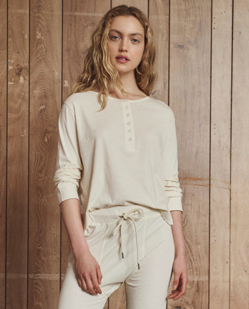 A person with long, wavy blonde hair wears THE GREAT. H25 SLEEP's The Crop Sleep Henley in Washed White, paired with matching cotton drawstring pants, standing before a wooden plank wall.