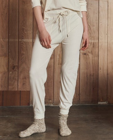A person stands indoors in THE GREAT. H25 SLEEP The Long John. -- Washed White, paired with a matching top and gray speckled socks, against a wooden wall.