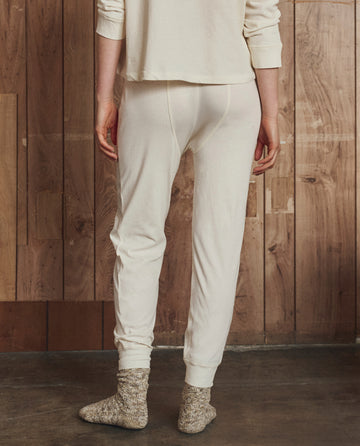 A person stands indoors facing a wooden wall, wearing THE GREAT. H25 SLEEP The Long John in Washed White and marled gray socks; only the lower half is visible.